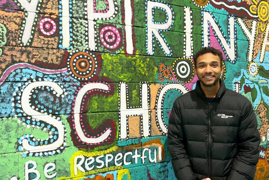 An aboriginal man standing in front of a colorful wall with the inscriptions Yprinia School